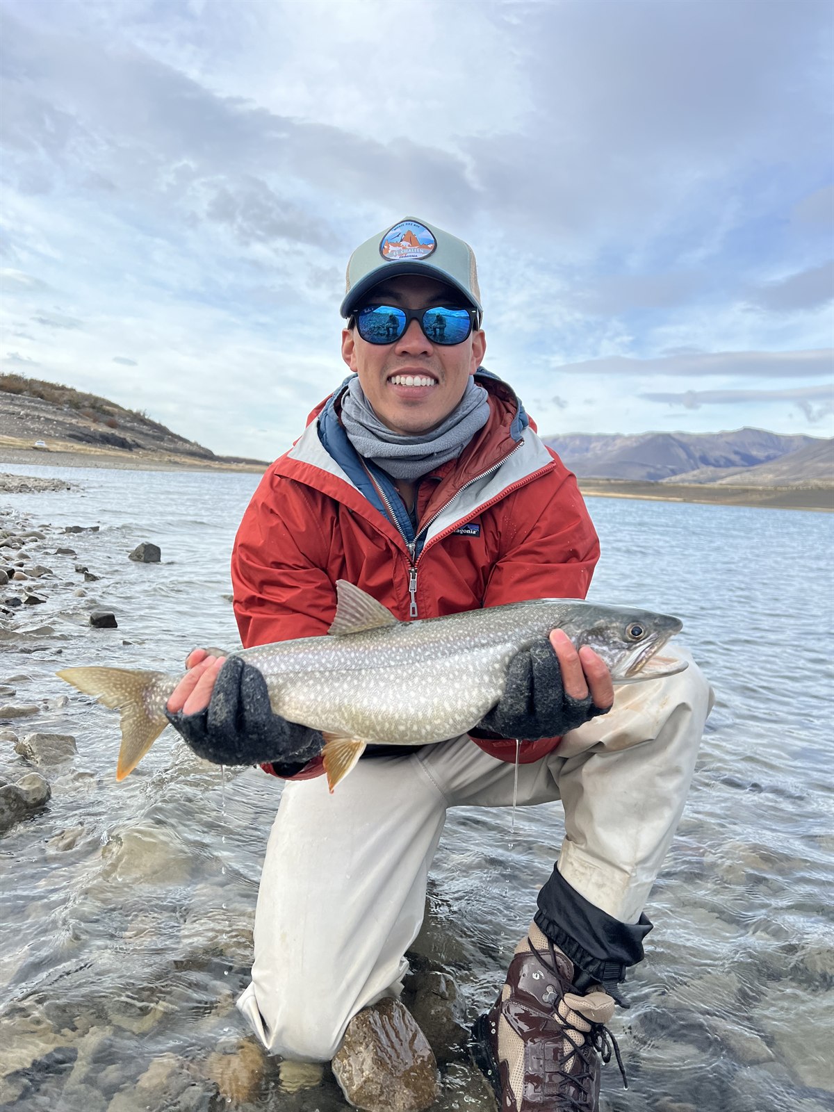 Phong Nguyen kneeling in a river in a red jacket, smiling and holding a large spotted fish